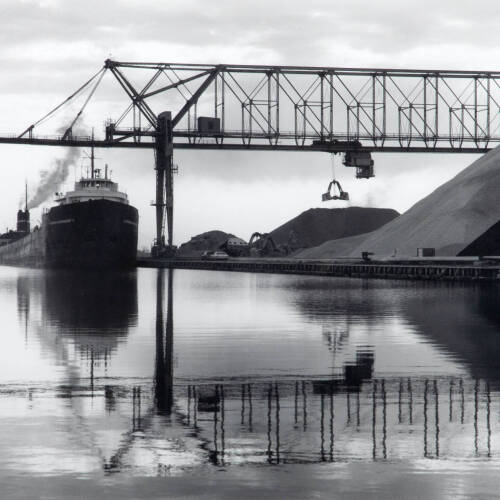 Black and white photograph of a steam ship in water, docked; reflections rippling in water.